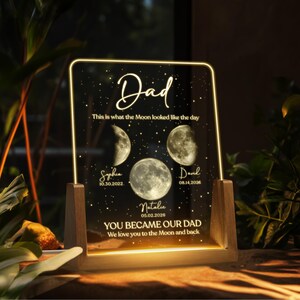 K&ouml;nnte beinhalten: Ein personalisiertes Acryl-Nachtlicht mit Holzsockel. Die transparente Acrylplatte wird mit warmem Licht beleuchtet und zeigt das Wort "Dad" in eleganter Schrift, zusammen mit Mondphasen-Grafiken und Namen mit Daten. Der Text lautet "YOU BECAME OUR DAD We love you to the Moon and back."