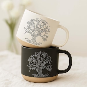 May include: Two ceramic mugs stacked vertically. The top mug is off-white with a black line-art tree design featuring a heart with the text S+J. The bottom mug is matte black with a white line-art tree design featuring a heart with the text S+J.