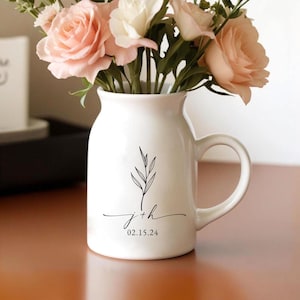 May include: A white ceramic vase with a handle, filled with pink and white flowers. The vase has a black leaf design and the initials "j+h" with the date "02.15.24" printed below. The vase is on a wooden surface.