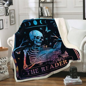 Puede incluir: Una manta de tiro negra y azul con un esqueleto sosteniendo un libro. El libro tiene un dise&ntilde;o de galaxia y el texto "The Reader" est&aacute; impreso en la manta. La manta tiene una textura blanca y esponjosa.