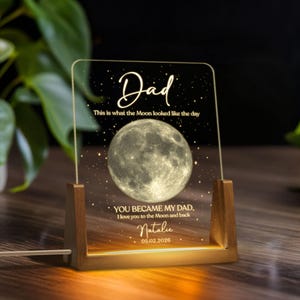 K&ouml;nnte beinhalten: Personalisiertes Nachtlicht mit Holzsockel und klarer Acrylplatte. Die Platte zeigt das Wort "Dad" und ein detailliertes Mondbild sowie einen individuellen Text und ein Datum. Das Licht strahlt in einem warmen, einladenden Farbton.