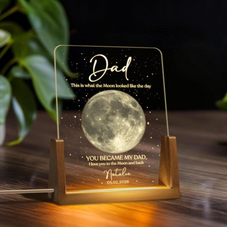 K&ouml;nnte beinhalten: Personalisiertes Nachtlicht mit Holzsockel und klarer Acrylplatte. Die Platte zeigt das Wort "Dad" und ein detailliertes Mondbild sowie einen individuellen Text und ein Datum. Das Licht strahlt in einem warmen, einladenden Farbton.