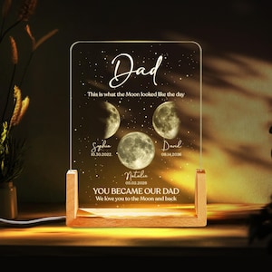 K&ouml;nnte beinhalten: Ein personalisiertes Nachtlicht mit Holzsockel und klarer Acrylplatte. Die Platte zeigt das Wort "Dad" in Schrift, Mondphasenillustrationen, Namen und Daten. Der Text enth&auml;lt "You became our Dad" und "We love you to the Moon and back."