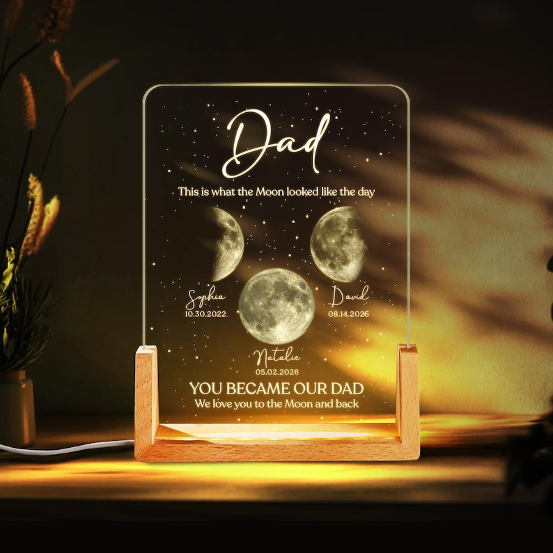 K&ouml;nnte beinhalten: Ein personalisiertes Nachtlicht mit Holzsockel und klarer Acrylplatte. Die Platte zeigt das Wort "Dad" in Schrift, Mondphasenillustrationen, Namen und Daten. Der Text enth&auml;lt "You became our Dad" und "We love you to the Moon and back."