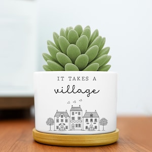 May include: A white ceramic planter with a light wood saucer base containing a green succulent. The planter features black text that reads IT TAKES A village above a line drawing of three houses, two trees, and three small birds in flight.