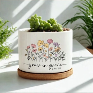 May include: A white ceramic planter with a succulent plant. The planter features a floral design in pastel colors and the text "grow in grace" with a Bible verse. The planter sits on a round, wooden saucer.