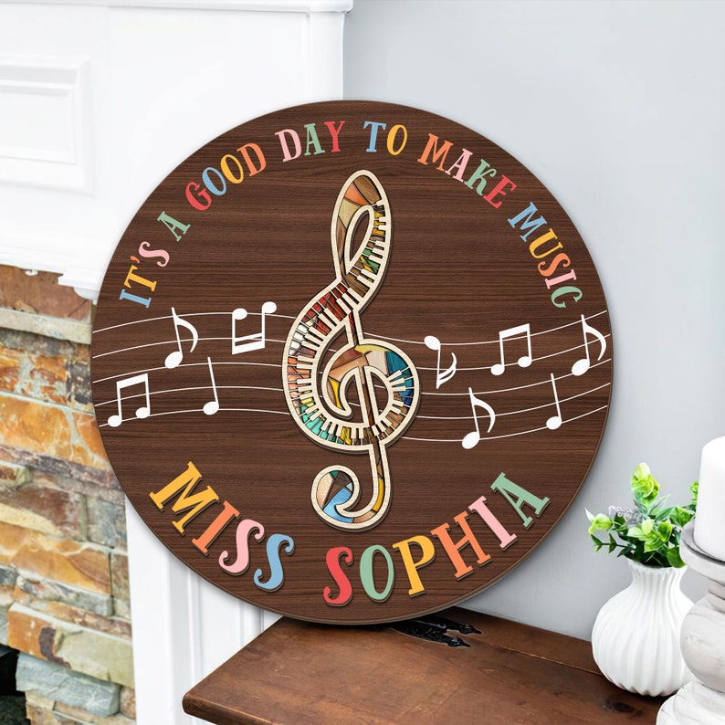 May include: A round wooden sign with a brown background and a colorful treble clef in the center. The sign says "It's a good day to make music" in rainbow letters around the treble clef. The words "Miss Sophia" are written in rainbow letters at the bottom of the sign.