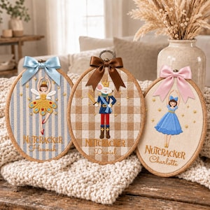 May include: Three oval wooden embroidery hoops featuring Nutcracker-themed designs. The left hoop has a blue striped fabric with a fairy, the centre has tan gingham with a mouse soldier, and the right has beige fabric with a ballerina, all with bows.