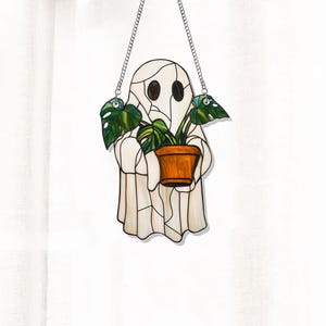 May include: A stained glass ghost holding a potted plant with green leaves. The ghost is white with a textured surface and has black eyes. The pot is brown and the plant is green.