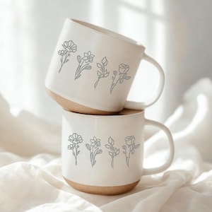 May include: Two white ceramic mugs stacked on top of each other. Each mug has a light brown base and a white handle. The mugs are decorated with a gray floral line drawing. The mugs are sitting on a white cloth.