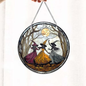 May include: A stained glass window hanging with a silver chain. The window depicts three witches dancing in a forest under a full moon. The witches are wearing black hats and long dresses in shades of purple, yellow, and gray.