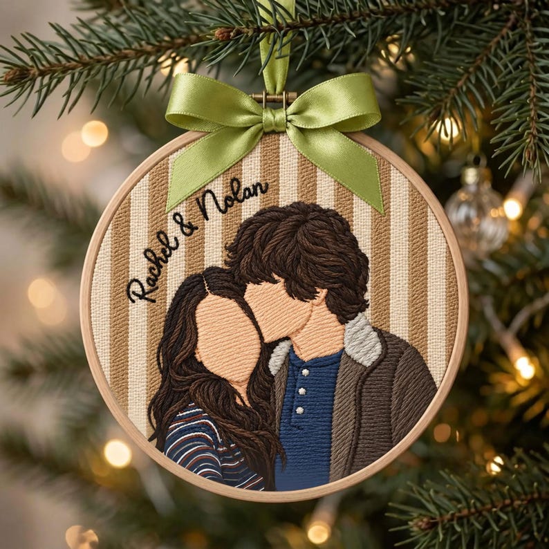 May include: A circular embroidery hoop ornament featuring a stitched portrait of two people with brown hair, one wearing a blue shirt and brown jacket, the other a striped top. The text "Rachel & Nolan" is embroidered above, topped with a green ribbon.