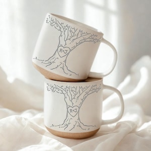 May include: Two white ceramic mugs stacked, each with a grey tree design and initials "R+J" in a heart. The mugs have a natural, light brown base and a curved handle.