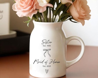 May include: White ceramic vase with a handle, featuring the text "Sister Est. 1995" and "Maid of Honor Est. 2025" with a heart symbol.