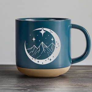 K&ouml;nnte beinhalten: Eine tealblaue Keramiktasse mit einem wei&szlig;en Design aus Bergen, einer Mondsichel und Sternen. Die Tasse hat einen beigefarbenen Boden.