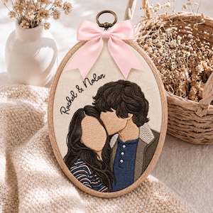 May include: An oval embroidery hoop featuring a custom thread portrait of two people with the text Rachel & Nolan stitched in black. The hoop is adorned with a light pink satin ribbon bow at the top and a bronze-colored metal hanging loop.