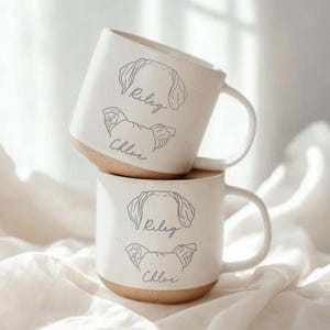 May include: Two white ceramic mugs stacked on top of each other. Each mug has a light brown base and a handle. The mugs are decorated with line drawings of dog ears and the names "Riley" and "Chloe" in cursive.