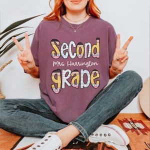 以下が含まれることがあります： 「Second Grade」の文字が鉛筆風のアクセントで遊び心のあるフォントでプリントされたマルーン色のTシャツ。「Mrs. Harrington」の文字が「Second」の下にプリントされています。快適なフィット感のシャツです。