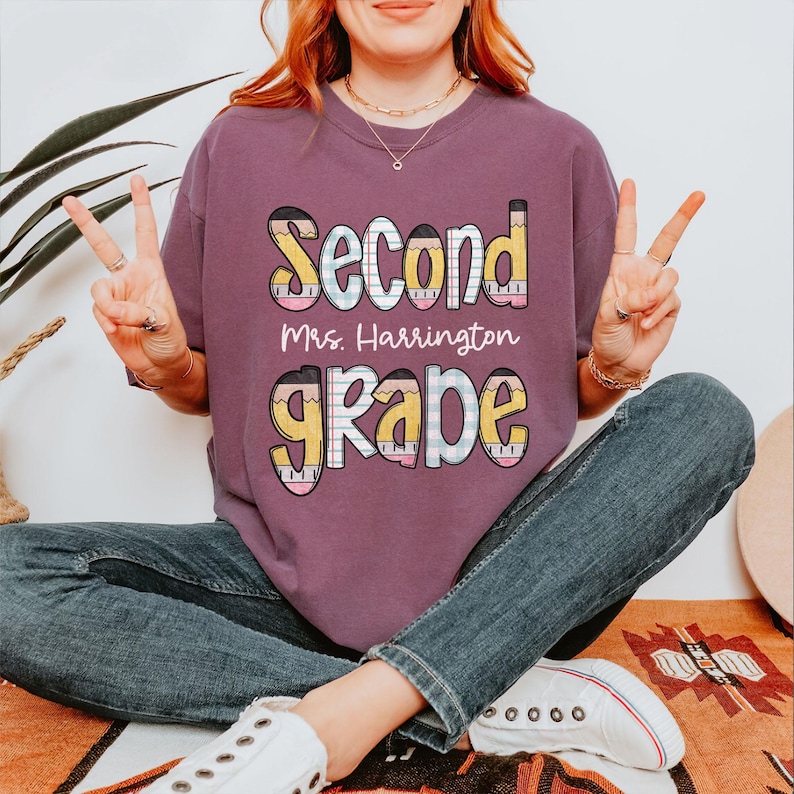 以下が含まれることがあります： 「Second Grade」の文字が鉛筆風のアクセントで遊び心のあるフォントでプリントされたマルーン色のTシャツ。「Mrs. Harrington」の文字が「Second」の下にプリントされています。快適なフィット感のシャツです。