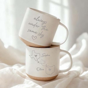 May include: Two white ceramic mugs stacked on top of each other. The top mug has the text "always under the same sky" and a heart. The bottom mug features a map of Texas and Ohio with the names "Sophia" and "Olivia".