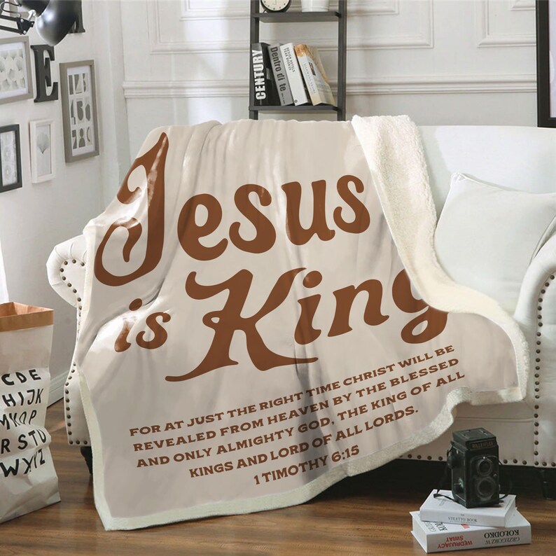 Może przedstawiać: Kremowy koc z napisem "Jesus is King" w brązowym skrypcie. Koc ma podszewkę z sherpy i werset biblijny wydrukowany na dole. Koc jest narzucony na białą sofę.