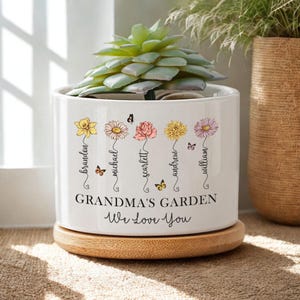 May include: White ceramic planter with a succulent plant. The planter features floral illustrations with names and the text "GRANDMA'S GARDEN We Love You". It sits on a round wooden base. Butterflies accent the floral design.