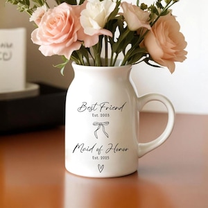 May include: White ceramic vase with a handle, filled with pink and white flowers. The vase has the text "Best Friend Est. 2003" and "Maid of Honor Est. 2025" with a bow and heart graphic.