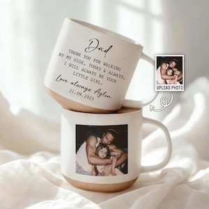 May include: Two white ceramic mugs stacked on top of each other. The top mug has the word "Dad" and a message printed on it. The bottom mug has a photo of a man and a child. The mugs have a tan base.