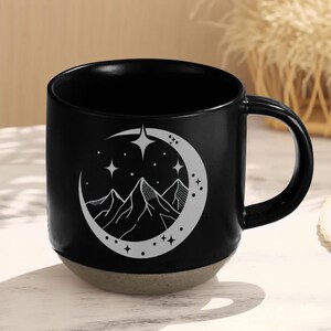 K&ouml;nnte beinhalten: Schwarze Keramiktasse mit einem Design aus Halbmond und Bergen. Der Mond ist wei&szlig; umrandet und hat Sterne im Inneren. Die Berge sind ebenfalls wei&szlig; und befinden sich innerhalb des Mondes.