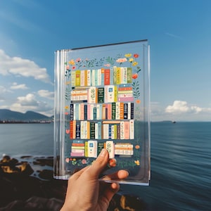 May include: A clear acrylic notebook with a colorful illustration of a bookshelf filled with books and flowers. The notebook is being held in a hand against a blue sky and ocean background.