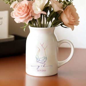 May include: White ceramic vase with a handle, featuring a rainbow outline of two hands holding each other. The vase is filled with pink and white roses. The bottom of the vase has the text "y + h" and the date "02.11.2025" in a purple color.