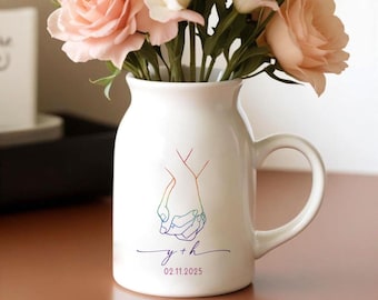 May include: White ceramic vase with a handle, featuring a rainbow outline of two hands holding each other. The vase is filled with pink and white roses. The bottom of the vase has the text "y + h" and the date "02.11.2025" in a purple color.