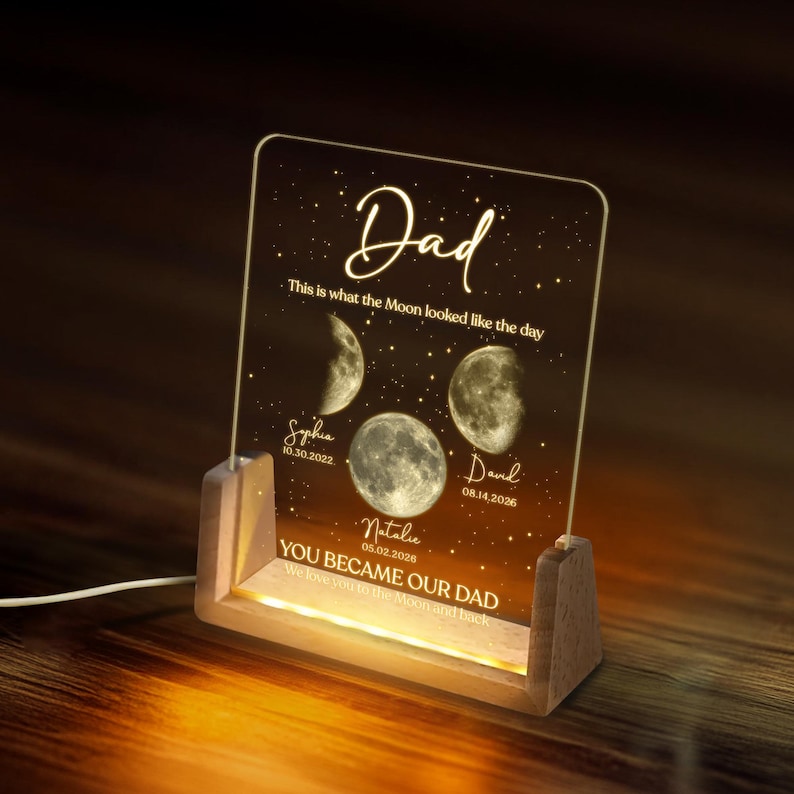 K&ouml;nnte beinhalten: Ein personalisiertes Acryl-Nachtlicht mit Holzsockel. Das Licht zeigt die Mondphasen und die Namen und Daten der Lieben an. Der Text lautet "Papa" und "Du wurdest unser Papa. Wir lieben dich bis zum Mond und zur&uuml;ck."