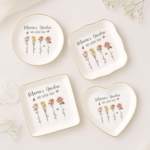 Puede incluir: Cuatro platos de cer&aacute;mica blanca con borde dorado para guardar peque&ntilde;os objetos. Cada plato tiene un dise&ntilde;o floral con mariposas y el texto "Mama's Garden 4 We Love You". Los nombres "Charlea", "Maggie", "Hayden" y "Aubrey" est&aacute;n escritos debajo de las flores.