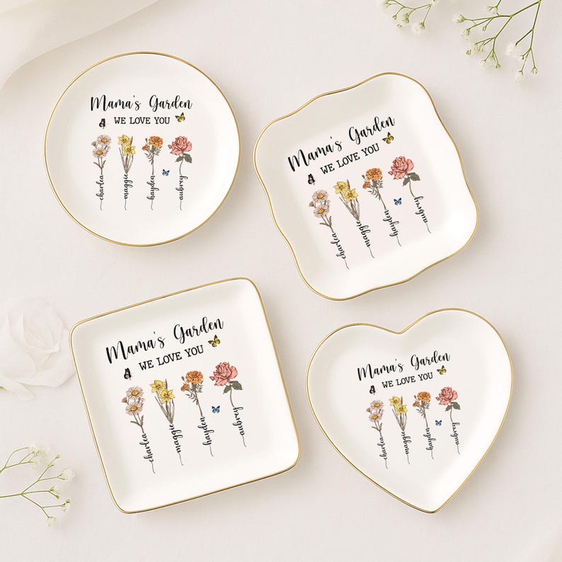 Puede incluir: Cuatro platos de cer&aacute;mica blanca con borde dorado para guardar peque&ntilde;os objetos. Cada plato tiene un dise&ntilde;o floral con mariposas y el texto "Mama's Garden 4 We Love You". Los nombres "Charlea", "Maggie", "Hayden" y "Aubrey" est&aacute;n escritos debajo de las flores.