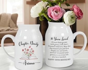 May include: Two white ceramic mugs with handles, one with "Chapter Ninety" and "Ariana" printed on it, and the other with "90 Years Loved" text. A vase with pink and white flowers sits behind the mugs.