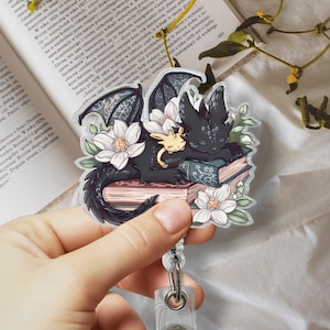 May include: A black and white dragon and a smaller yellow dragon are curled up asleep on top of a stack of books. The dragons are surrounded by white flowers and green leaves. The image is a sticker.