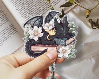 Puede incluir: Un dragón negro y blanco y un dragón amarillo más pequeño están acurrucados y dormidos encima de una pila de libros. Los dragones están rodeados de flores blancas y hojas verdes. La imagen es una pegatina.