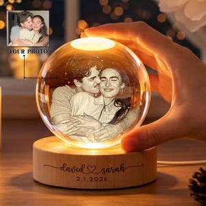 May include: A personalized crystal ball with a photo of a couple, illuminated by a warm light. The wooden base is engraved with the names "David & Sarah" and the date "2.1.2026". The words "YOUR PHOTO" are visible in the image.