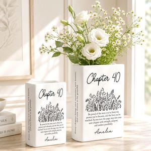 May include: Two white rectangular ceramic vases featuring the black text Chapter 40, a floral line drawing, the name Amelia, and a quote about new chapters. The taller vase holds a bouquet of white roses and small white wildflowers.