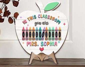 May include: Wooden apple-shaped sign with the text "IN THIS CLASSROOM you are MRS. SOPHIA" and colorful crayon illustrations. The sign is white with a wooden stand, and the apple has a green leaf. The sign is designed for a teacher.