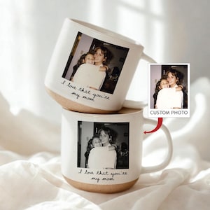May include: Two white ceramic mugs with a photo of a mother and child. The mugs have a tan base and the text "I love that you're my mom" in black script. A small photo with the words "CUSTOM PHOTO" is also shown.