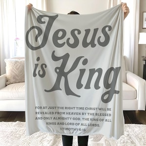 Może przedstawiać: Jasnoszary koc z napisem "Jesus is King" dużymi, stylizowanymi szarymi literami. Poniżej znajduje się cytat z 1 Tymoteusza 6:15 mniejszym szarym tekstem. Koc jest prezentowany przed białą sofą.