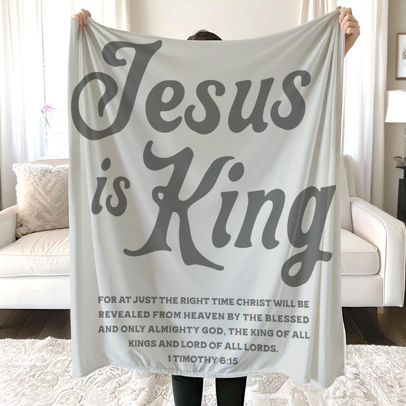 Może przedstawiać: Jasnoszary koc z napisem "Jesus is King" dużymi, stylizowanymi szarymi literami. Poniżej znajduje się cytat z 1 Tymoteusza 6:15 mniejszym szarym tekstem. Koc jest prezentowany przed białą sofą.