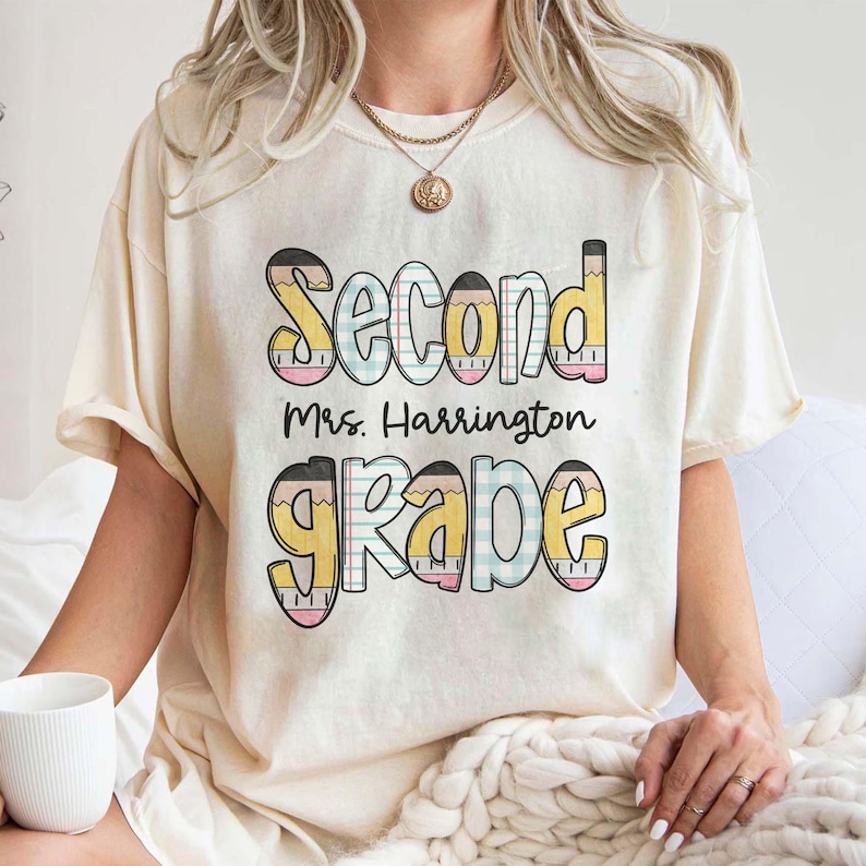 以下が含まれることがあります： クリーム色のTシャツ。カラフルな鉛筆をテーマにしたデザインで「Second Mrs. Harrington Grade」の文字がプリントされています。「Second」は青色、「Grade」は黄色で、遊び心のあるフォントで書かれています。