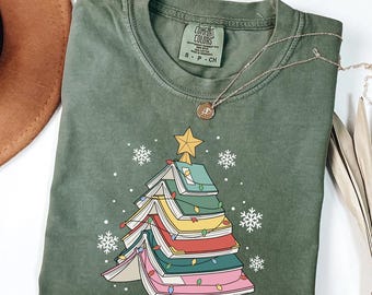 May include: Green Comfort Colors t-shirt featuring a Christmas tree design made of colorful books, adorned with a gold star and string lights. White snowflakes are scattered around the tree. A gold necklace is draped across the shirt.