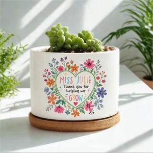 May include: A white ceramic planter with a succulent plant. The planter has a heart-shaped floral design with the text "Miss Julie, Thank you for helping me GROW". The planter sits on a round, wooden coaster.