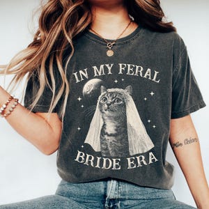 May include: A dark gray t-shirt with a graphic of a cat wearing a veil and the text "In My Feral Bride Era".