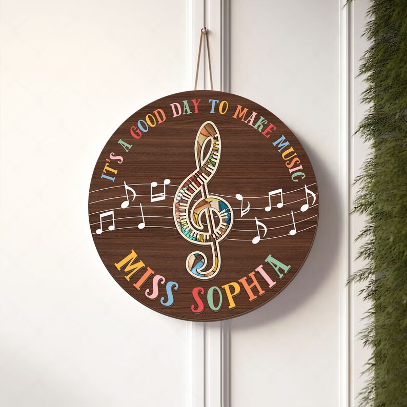 May include: A round wooden sign with a brown background and a colorful treble clef in the center. The sign says "It's a good day to make music" in a rainbow font around the treble clef. The words "Miss Sophia" are written in a rainbow font at the bottom of the sign.