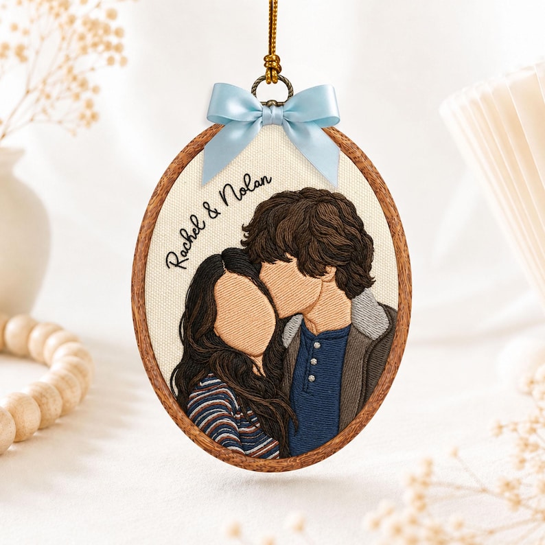 May include: An oval-shaped embroidery hoop ornament featuring a thread-stitched portrait of two people. The text "Rachel & Nolan" is embroidered in black. The hoop is framed in wood and topped with a light blue satin ribbon bow for hanging.